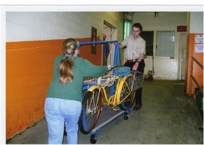 ellow bike on blue cart being wheeled in to a hallway with yellow walls.  A woman pushes from one side, stabilizing the bike with her hand. A man pulls form the other side. The bike has baskets in front and back.